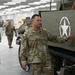 A group of U.S. Army soldiers in MultiCam (OCP) uniforms are touring what appears to be a military museum or exhibit hall. The central figure, whose name tape reads 'ABELLA,' is standing next to a vintage WWII-era half-track vehicle bearing the white Allied star insignia. The setting is an indoor facility with modern lighting, suggesting an organized tour or official visit.