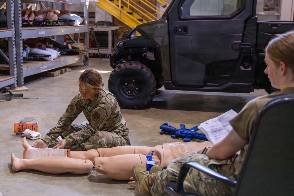 Two military personnel in camouflage uniforms are conducting a combat casualty care (TCCC) training exercise inside a warehouse or hangar facility. One service member is kneeling on the floor applying bandaging techniques to a medical training mannequin, while a second observes and reviews documents. Training props including a blue training rifle and SAM splint packaging are visible nearby.