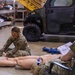 Two military personnel in camouflage uniforms are conducting a combat casualty care (TCCC) training exercise inside a warehouse or hangar facility. One service member is kneeling on the floor applying bandaging techniques to a medical training mannequin, while a second observes and reviews documents. Training props including a blue training rifle and SAM splint packaging are visible nearby.