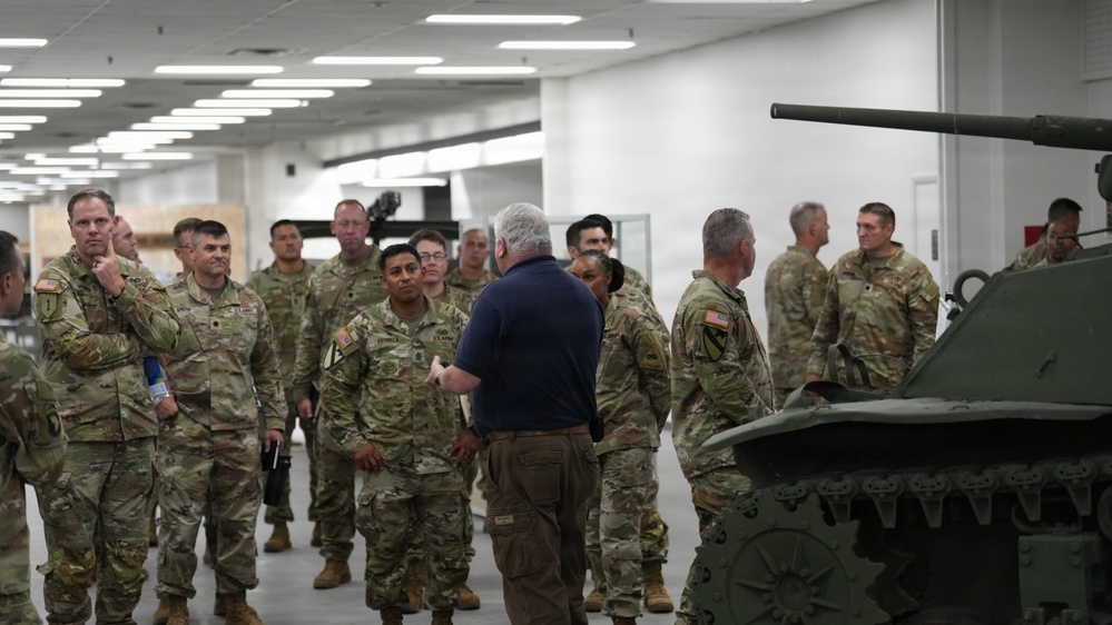 A group of U.S. Army soldiers in OCP (Operational Camouflage Pattern) uniforms are gathered in what appears to be an indoor facility or exhibition hall. A civilian in a navy polo shirt and khaki pants appears to be briefing or presenting to the assembled military personnel. A green military vehicle, likely a robotic or unmanned ground vehicle with a turret, is visible in the foreground right.
