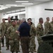 A group of U.S. Army soldiers in OCP (Operational Camouflage Pattern) uniforms are gathered in what appears to be an indoor facility or exhibition hall. A civilian in a navy polo shirt and khaki pants appears to be briefing or presenting to the assembled military personnel. A green military vehicle, likely a robotic or unmanned ground vehicle with a turret, is visible in the foreground right.