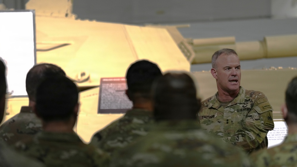 A U.S. Army officer with the name tape 'TAYLOR' and visible rank insignia (two stars, indicating a Major General) is addressing a group of soldiers in camouflage uniforms. The briefing or presentation appears to be taking place in a facility containing military equipment, specifically what appears to be a tan/desert-colored armored vehicle or tank in the background. The setting suggests a formal military briefing or orientation event.