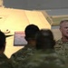 A U.S. Army officer with the name tape 'TAYLOR' and visible rank insignia (two stars, indicating a Major General) is addressing a group of soldiers in camouflage uniforms. The briefing or presentation appears to be taking place in a facility containing military equipment, specifically what appears to be a tan/desert-colored armored vehicle or tank in the background. The setting suggests a formal military briefing or orientation event.