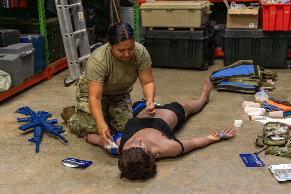 A female soldier in military camouflage (OCP/MultiCam) uniform is conducting combat casualty care training on a medical mannequin laid on a warehouse floor. The training scenario simulates treating a wounded casualty, with medical supplies and training weapons visible around the scene. The setting appears to be a military storage facility or training area.