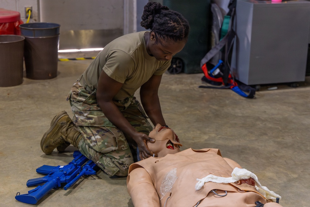 A soldier in U.S. Army OCP (Operational Camouflage Pattern) uniform is kneeling on a concrete floor performing combat casualty care training on a medical simulation mannequin. The mannequin features realistic wound simulation including simulated facial and chest injuries. Blue training rifles are visible nearby, indicating this is a Tactical Combat Casualty Care (TCCC) or similar military medical training exercise.