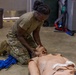 A soldier in U.S. Army OCP (Operational Camouflage Pattern) uniform is kneeling on a concrete floor performing combat casualty care training on a medical simulation mannequin. The mannequin features realistic wound simulation including simulated facial and chest injuries. Blue training rifles are visible nearby, indicating this is a Tactical Combat Casualty Care (TCCC) or similar military medical training exercise.