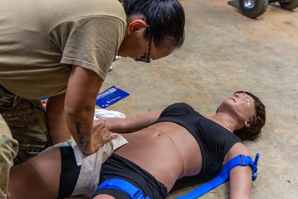 A military service member in camouflage uniform is performing first aid training on a realistic medical mannequin lying on a concrete floor. The trainee is applying wound packing or pressure bandaging to the mannequin's hip/groin area, which also has a blue tourniquet applied to the leg. This appears to be a Tactical Combat Casualty Care (TCCC) or similar military medical training exercise.