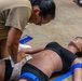 A military service member in camouflage uniform is performing first aid training on a realistic medical mannequin lying on a concrete floor. The trainee is applying wound packing or pressure bandaging to the mannequin's hip/groin area, which also has a blue tourniquet applied to the leg. This appears to be a Tactical Combat Casualty Care (TCCC) or similar military medical training exercise.