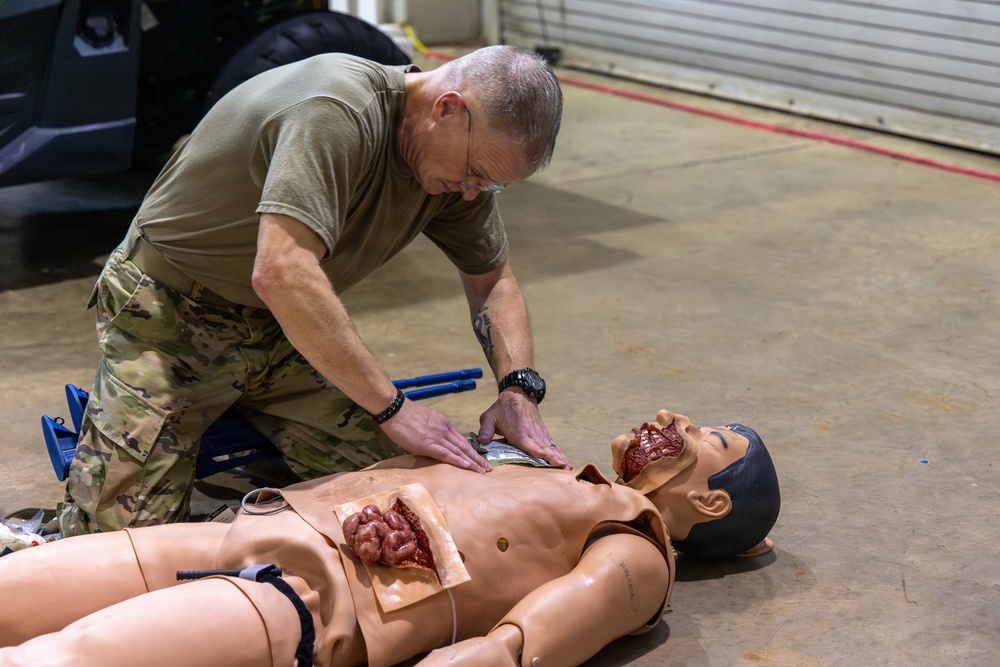 A military personnel member in camouflage uniform is conducting combat casualty care training using a realistic medical simulation mannequin. The mannequin features simulated traumatic wounds including abdominal and facial injuries, designed to replicate battlefield trauma scenarios. The training appears to take place in an indoor facility such as a hangar or training bay.