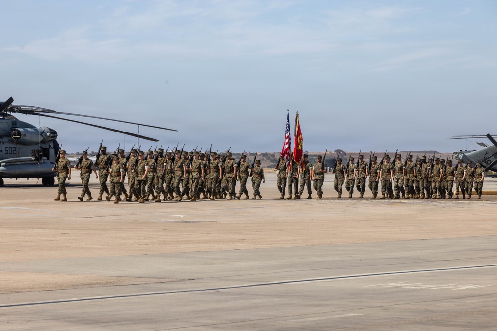 A large formation of U.S. Marines in woodland MARPAT camouflage uniforms conducts a formal parade or ceremony on a military airfield tarmac. The formation carries rifles at shoulder arms and is led by a color guard bearing the American flag and what appears to be a Marine Corps unit guidon. Military helicopters, including what appears to be a CH-53 on the left, are visible flanking the formation.