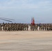 A large formation of U.S. Marines in woodland MARPAT camouflage uniforms conducts a formal parade or ceremony on a military airfield tarmac. The formation carries rifles at shoulder arms and is led by a color guard bearing the American flag and what appears to be a Marine Corps unit guidon. Military helicopters, including what appears to be a CH-53 on the left, are visible flanking the formation.