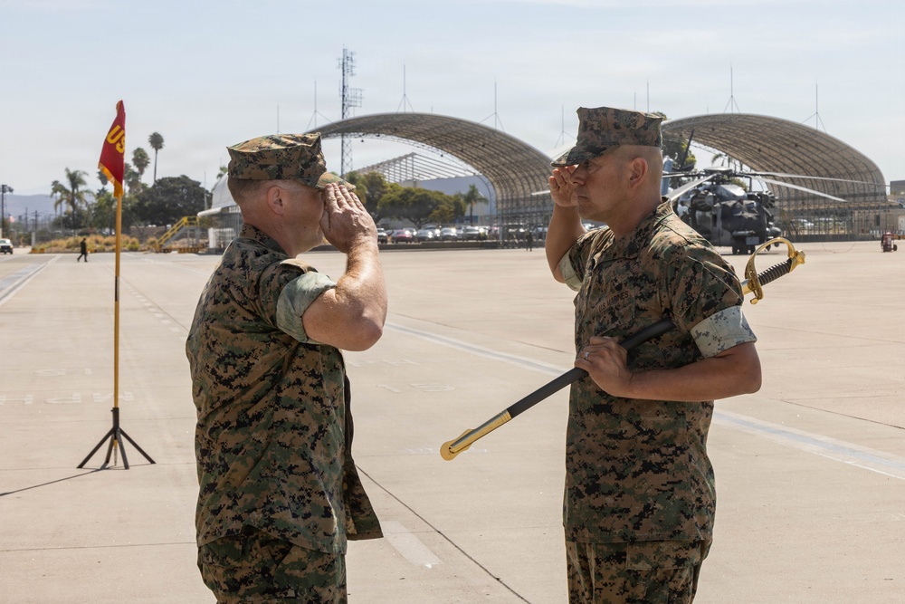 Two U.S. Marine Corps personnel in MARPAT digital camouflage uniforms are exchanging salutes on a military airfield tarmac. One Marine is holding a ceremonial sword, suggesting this is a formal change of command or military ceremony. The setting appears to be a Marine Corps Air Station, with aircraft hangars and a helicopter visible in the background.