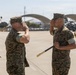 Two U.S. Marine Corps personnel in MARPAT digital camouflage uniforms are exchanging salutes on a military airfield tarmac. One Marine is holding a ceremonial sword, suggesting this is a formal change of command or military ceremony. The setting appears to be a Marine Corps Air Station, with aircraft hangars and a helicopter visible in the background.