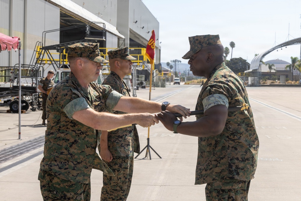 Three U.S. Marine Corps personnel in MARPAT woodland camouflage uniforms are participating in what appears to be a formal military ceremony on an airfield tarmac. Two Marines are exchanging or passing a ceremonial object, consistent with a change of command or non-commissioned officer sword ceremony. The setting appears to be a military air station, likely on the West Coast based on the visible palm trees and architecture.