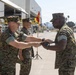 Three U.S. Marine Corps personnel in MARPAT woodland camouflage uniforms are participating in what appears to be a formal military ceremony on an airfield tarmac. Two Marines are exchanging or passing a ceremonial object, consistent with a change of command or non-commissioned officer sword ceremony. The setting appears to be a military air station, likely on the West Coast based on the visible palm trees and architecture.