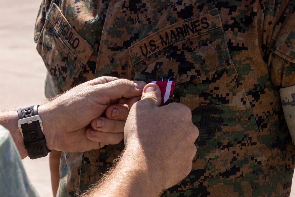 A close-up image showing hands pinning a medal onto a U.S. Marines uniform. The uniform displays the name tape 'ANGULO' on the left chest and 'U.S. MARINES' branch tape on the right, both in the standard MARPAT digital camouflage pattern. The medal being pinned appears to have a red and white ribbon, consistent with a military decoration ceremony.
