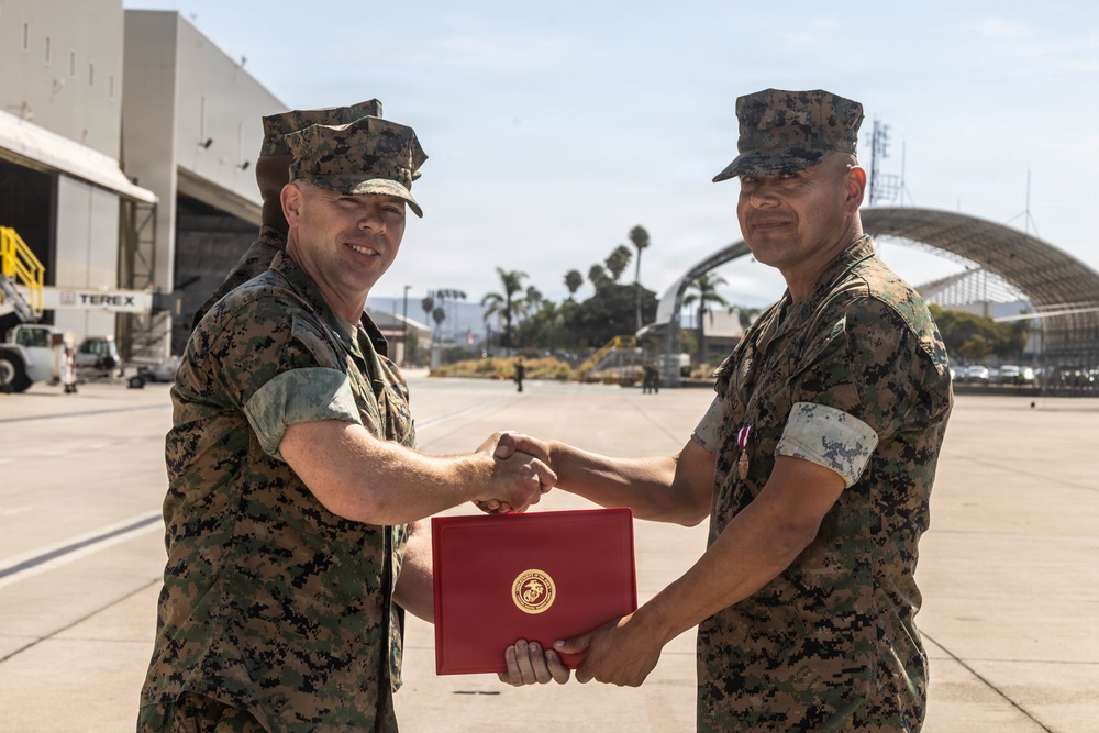 Two U.S. Marines in MARPAT digital camouflage uniforms are shaking hands while jointly holding a red presentation folder bearing the U.S. Marine Corps emblem (Eagle, Globe, and Anchor). The ceremony appears to be an award or commendation presentation taking place on a military airfield flight line. Large aircraft hangars, including one with an arched structure, and palm trees are visible in the background.