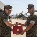 Two U.S. Marines in MARPAT digital camouflage uniforms are shaking hands while jointly holding a red presentation folder bearing the U.S. Marine Corps emblem (Eagle, Globe, and Anchor). The ceremony appears to be an award or commendation presentation taking place on a military airfield flight line. Large aircraft hangars, including one with an arched structure, and palm trees are visible in the background.