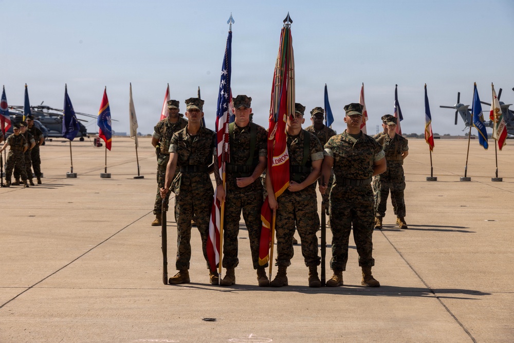A U.S. Marine Corps color guard stands at attention on a military airfield tarmac during a formal ceremony. The central group holds the American flag and what appears to be a Marine Corps unit guidon, surrounded by rows of state and unit flags. Military helicopters, including what appears to be CH-53s and V-22 Ospreys, are visible in the background.