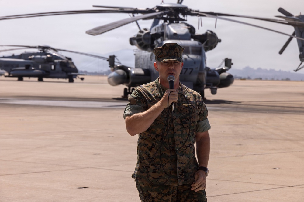 A U.S. Marine Corps officer in MARPAT camouflage uniform is speaking into a microphone on a military airfield tarmac. Large military helicopters, identifiable as CH-53E Super Stallions, are visible in the background. The setting appears to be an official ceremony or briefing conducted outdoors on the flight line.