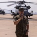 A U.S. Marine Corps officer in MARPAT camouflage uniform is speaking into a microphone on a military airfield tarmac. Large military helicopters, identifiable as CH-53E Super Stallions, are visible in the background. The setting appears to be an official ceremony or briefing conducted outdoors on the flight line.