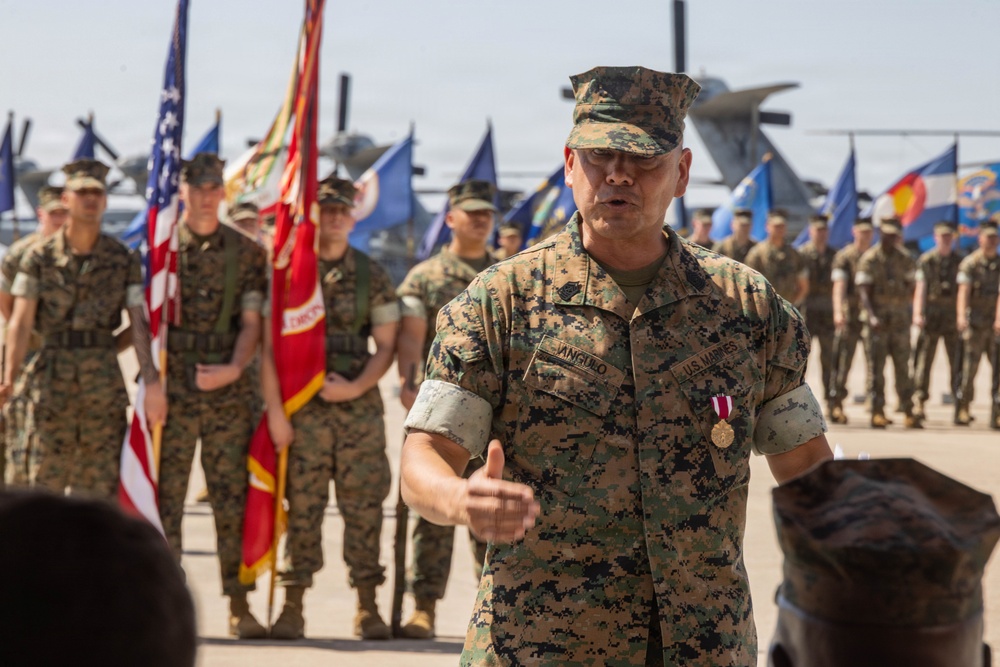 A U.S. Marine Corps officer named Angulo, wearing MARPAT camouflage utilities and displaying a medal on his chest, addresses a formation during what appears to be a military ceremony. He is speaking to an assembled group of Marines standing at parade rest behind him, with color guard personnel holding the American flag and unit guidons. The setting appears to be an outdoor tarmac or flight line, with military aircraft visible in the background.
