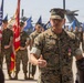 A U.S. Marine Corps officer named Angulo, wearing MARPAT camouflage utilities and displaying a medal on his chest, addresses a formation during what appears to be a military ceremony. He is speaking to an assembled group of Marines standing at parade rest behind him, with color guard personnel holding the American flag and unit guidons. The setting appears to be an outdoor tarmac or flight line, with military aircraft visible in the background.