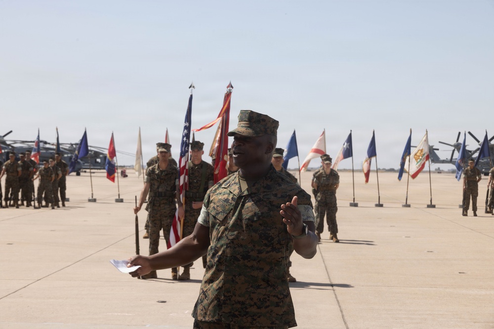 A U.S. Marine in digital camouflage (MARPAT) uniform is addressing a formation on a military flight line, holding papers and gesturing while speaking. Behind him, multiple color guard members hold unit guidons and the American flag, with rows of Marines standing at attention. Military helicopters are visible in the background, suggesting this is a formal ceremony at a Marine Corps air station.