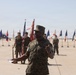 A U.S. Marine in digital camouflage (MARPAT) uniform is addressing a formation on a military flight line, holding papers and gesturing while speaking. Behind him, multiple color guard members hold unit guidons and the American flag, with rows of Marines standing at attention. Military helicopters are visible in the background, suggesting this is a formal ceremony at a Marine Corps air station.