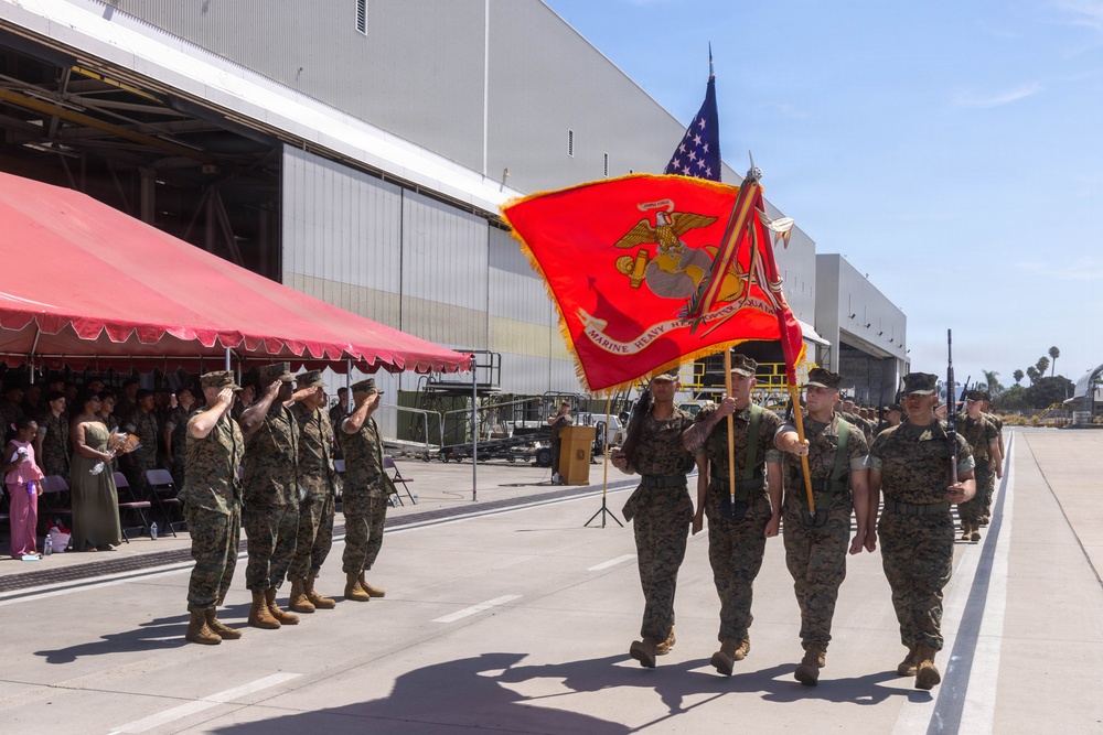 A United States Marine Corps color guard ceremony is taking place on a tarmac adjacent to large aircraft hangars. Marines in camouflage utilities are marching with the USMC flag and the American flag while fellow Marines render salutes. Civilians and family members are visible under a red canopy observing the formal military ceremony.