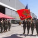 A United States Marine Corps color guard ceremony is taking place on a tarmac adjacent to large aircraft hangars. Marines in camouflage utilities are marching with the USMC flag and the American flag while fellow Marines render salutes. Civilians and family members are visible under a red canopy observing the formal military ceremony.