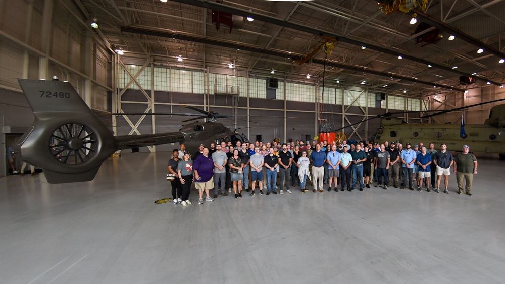 A large group of approximately 60-80 people pose for a group photograph inside a military aviation hangar. The hangar contains at least two military helicopters, including one prominently displayed with tail number 72480 in an olive/dark matte finish. The setting appears to be an official or commemorative gathering of military personnel and civilian workers.