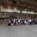 A large group of approximately 60-80 people pose for a group photograph inside a military aviation hangar. The hangar contains at least two military helicopters, including one prominently displayed with tail number 72480 in an olive/dark matte finish. The setting appears to be an official or commemorative gathering of military personnel and civilian workers.