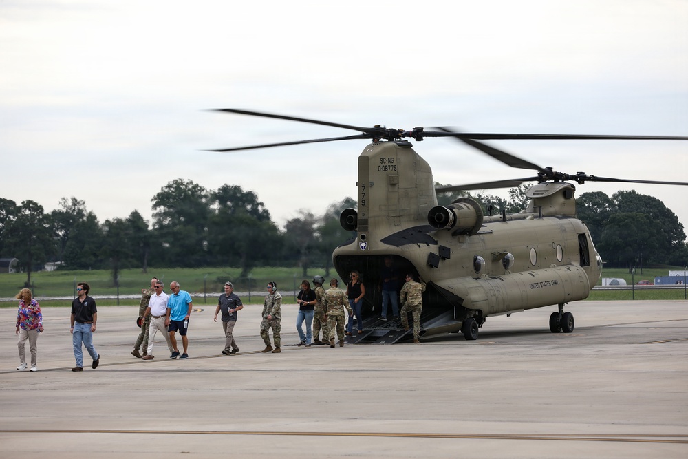 A United States Army CH-47 Chinook helicopter, marked with 'SC-NG' (South Carolina National Guard) and tail number D-08779, is parked on a tarmac with its rotors spinning. A mixed group of military personnel in camouflage uniforms and civilians in casual clothing are either disembarking or gathered near the rear loading ramp. The scene appears to be an official visit, tour, or dignitary transport operation at a military airfield.