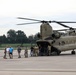A United States Army CH-47 Chinook helicopter, marked with 'SC-NG' (South Carolina National Guard) and tail number D-08779, is parked on a tarmac with its rotors spinning. A mixed group of military personnel in camouflage uniforms and civilians in casual clothing are either disembarking or gathered near the rear loading ramp. The scene appears to be an official visit, tour, or dignitary transport operation at a military airfield.