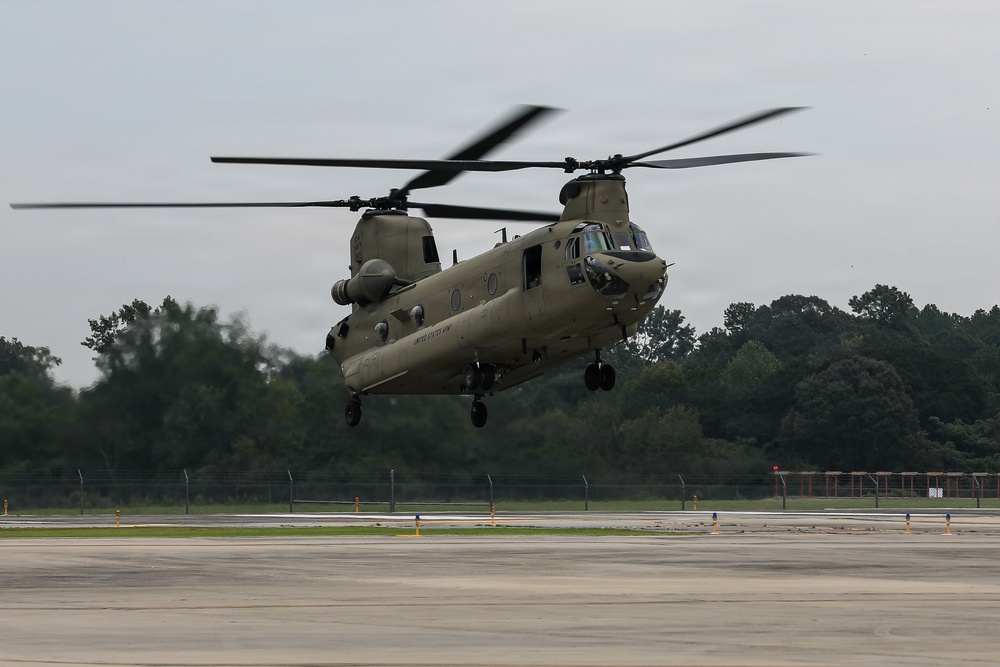 A U.S. Army CH-47 Chinook tandem-rotor helicopter is captured in low hover or takeoff/landing phase above a concrete tarmac. The aircraft is painted in standard olive drab military green and bears 'UNITED STATES ARMY' markings on its fuselage. The setting appears to be a military airfield surrounded by dense tree lines under overcast skies.