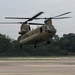 A U.S. Army CH-47 Chinook tandem-rotor helicopter is captured in low hover or takeoff/landing phase above a concrete tarmac. The aircraft is painted in standard olive drab military green and bears 'UNITED STATES ARMY' markings on its fuselage. The setting appears to be a military airfield surrounded by dense tree lines under overcast skies.