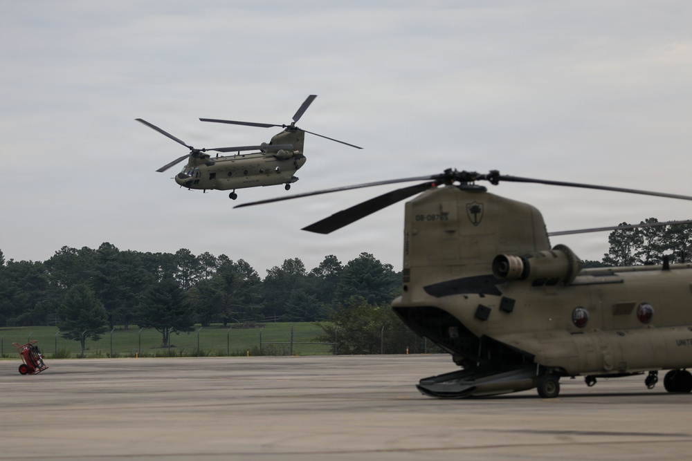 Two U.S. Army CH-47 Chinook tandem-rotor helicopters are visible on a military airfield. One Chinook is airborne and hovering or in low-level flight, while a second is parked on the tarmac in the foreground. The setting appears to be a military installation with a tree line and grassy areas visible in the background.