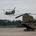 Two U.S. Army CH-47 Chinook tandem-rotor helicopters are visible on a military airfield. One Chinook is airborne and hovering or in low-level flight, while a second is parked on the tarmac in the foreground. The setting appears to be a military installation with a tree line and grassy areas visible in the background.