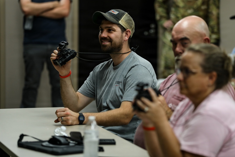 A group of civilians are seated at a table during what appears to be a demonstration or briefing, handling what look like night vision or optical devices. The central figure, a smiling man in a gray t-shirt and camouflage cap, holds up a binocular-style device on a neck lanyard. In the background, a person in military camouflage uniform is partially visible.