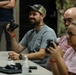 A group of civilians are seated at a table during what appears to be a demonstration or briefing, handling what look like night vision or optical devices. The central figure, a smiling man in a gray t-shirt and camouflage cap, holds up a binocular-style device on a neck lanyard. In the background, a person in military camouflage uniform is partially visible.