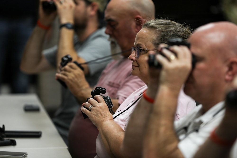 A group of adults seated in a row are holding cameras up to their eyes, appearing to be participating in a photography class or workshop. They are seated at a long table in what appears to be an indoor venue. Multiple participants are visible in profile, all engaged in the same activity of looking through their camera viewfinders.