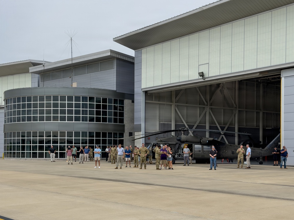 A group of approximately 30-40 individuals, including military personnel in camouflage uniforms and civilians, are gathered on an aircraft apron in front of a large hangar. Inside and near the hangar entrance, at least two military helicopters (appearing to be UH-60 Black Hawks) are visible. The setting appears to be a military aviation facility or National Guard installation, possibly during a ceremony, briefing, or tour.