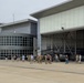 A group of approximately 30-40 individuals, including military personnel in camouflage uniforms and civilians, are gathered on an aircraft apron in front of a large hangar. Inside and near the hangar entrance, at least two military helicopters (appearing to be UH-60 Black Hawks) are visible. The setting appears to be a military aviation facility or National Guard installation, possibly during a ceremony, briefing, or tour.