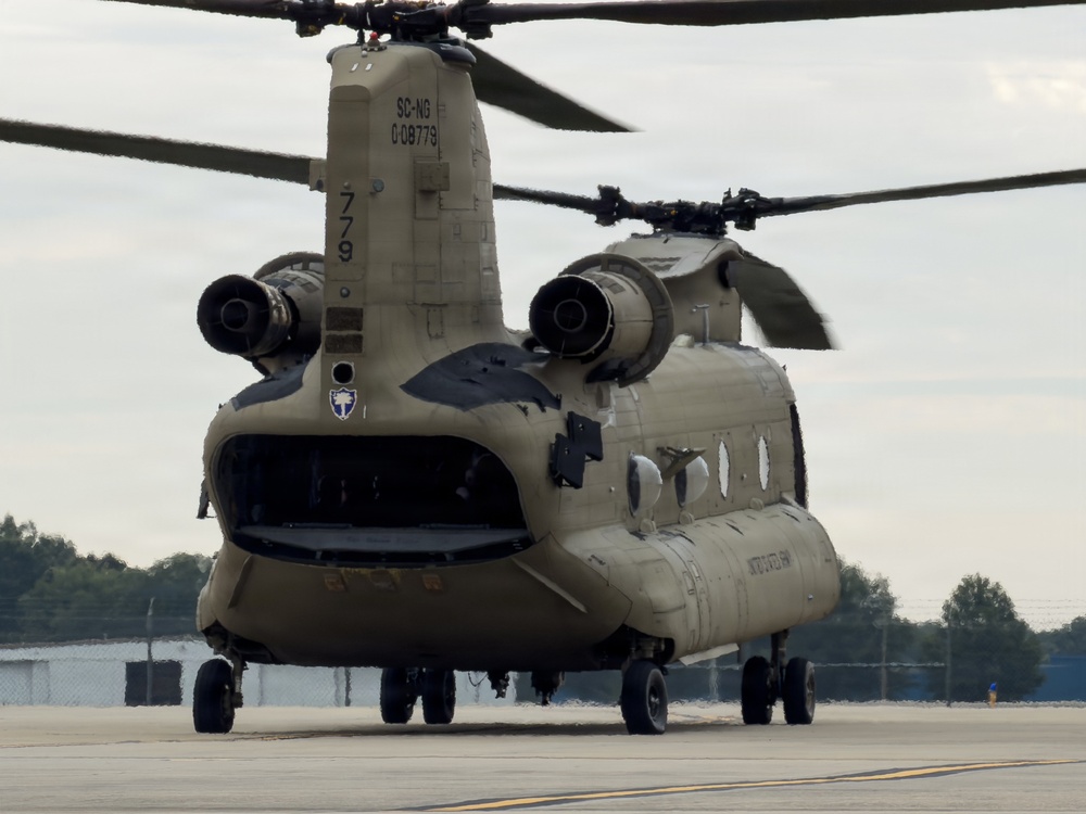 A U.S. Army CH-47 Chinook tandem-rotor helicopter is parked on a tarmac apron, identified by markings 'SC-NG 008779' indicating it belongs to the South Carolina National Guard. The aircraft is viewed from the front-rear quarter angle, showing both rotor systems, twin engines, and an open rear ramp. The rotors appear to be spinning, suggesting the aircraft is running or has recently been operating.