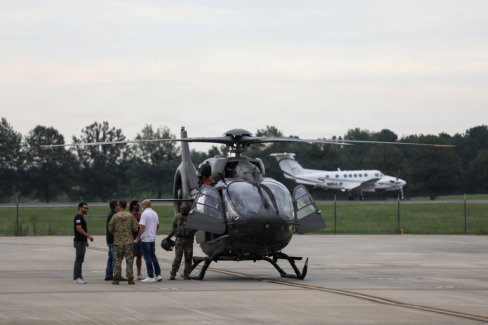 A group of approximately five to six individuals, including military personnel in camouflage uniforms and civilians, are gathered on an airport tarmac next to a dark gray military-style helicopter. The scene appears to be a briefing or post-flight discussion near the aircraft. In the background, a white twin-engine turboprop aircraft is visible on the runway.