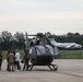A group of approximately five to six individuals, including military personnel in camouflage uniforms and civilians, are gathered on an airport tarmac next to a dark gray military-style helicopter. The scene appears to be a briefing or post-flight discussion near the aircraft. In the background, a white twin-engine turboprop aircraft is visible on the runway.