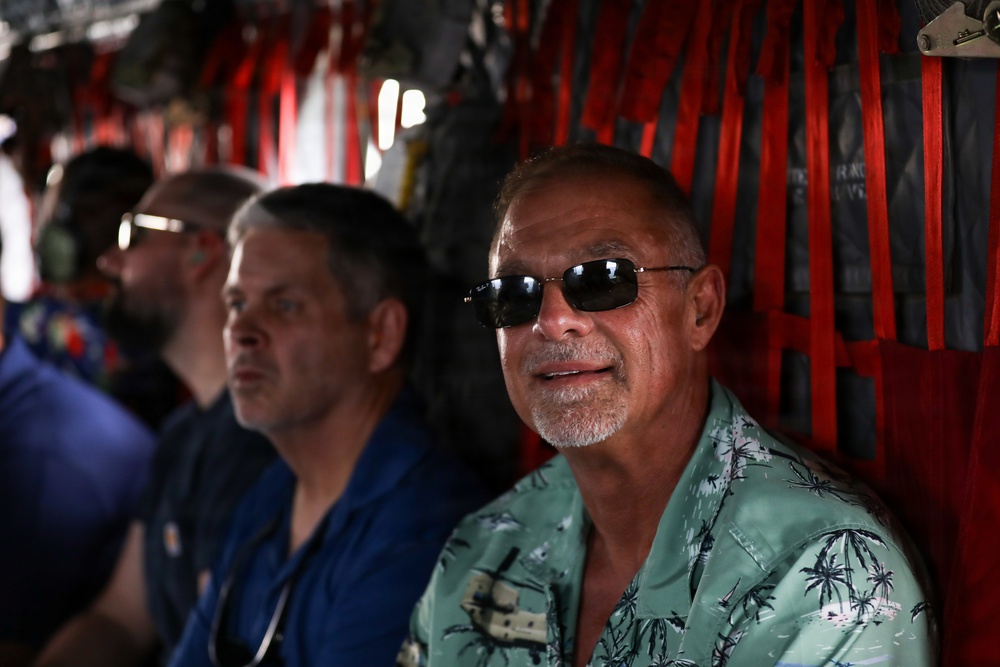 Several men are seated inside what appears to be a military cargo aircraft, identifiable by the red cargo straps and utilitarian interior. The foreground subject is a middle-aged man wearing a light green Hawaiian shirt and dark sunglasses, appearing relaxed. Behind him are at least two other men in blue shirts, slightly out of focus.