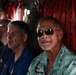 Several men are seated inside what appears to be a military cargo aircraft, identifiable by the red cargo straps and utilitarian interior. The foreground subject is a middle-aged man wearing a light green Hawaiian shirt and dark sunglasses, appearing relaxed. Behind him are at least two other men in blue shirts, slightly out of focus.