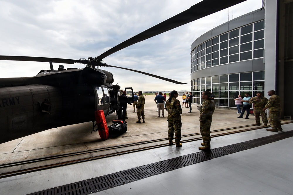 A U.S. Army UH-60 Black Hawk helicopter is parked on a tarmac adjacent to a modern curved glass building, likely a terminal or aviation facility. Military personnel in camouflage uniforms and civilians are gathered around the aircraft, suggesting a demonstration, open house, or familiarization event. The scene appears to be at a National Guard or military airfield facility.
